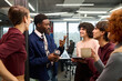 © TrueFrame Collective - Group of young adult multiethnic colleagues standing in modern office, engaging in lively conversation, Black man gesturing while holding reusable coffee cup, others listening and smiling