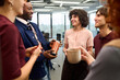 © TrueFrame Collective - Group of diverse young adults standing in modern office, smiling and talking during coffee break, multiethnic colleagues interacting and holding mugs, casual business meeting atmosphere