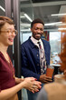 © TrueFrame Collective - Young adult Black man smiling while holding reusable coffee cup, standing with multiethnic group of young adults in modern office setting during casual conversation