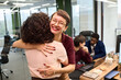 © TrueFrame Collective - Young adult Caucasian woman smiling and hugging young adult woman with curly hair in modern office, two young adult coworkers including Black man and Caucasian man working at laptops in background