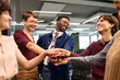 © TrueFrame Collective - Diverse group of young adults standing in modern office joining hands in center smiling and interacting with each other showing teamwork and collaboration during meeting