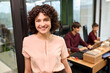© TrueFrame Collective - Portrait of young adult Caucasian woman smiling at camera in modern office, two young adult colleagues working together on laptops in background, open workspace environment visible