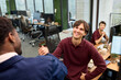 © TrueFrame Collective - Caucasian young adult man smiling while shaking hands with Black young adult man in modern office, young adult Caucasian woman sitting at desk in background observing interaction