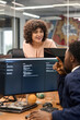 © TrueFrame Collective - Caucasian young adult woman smiling while talking to Black young adult man sitting at desk using computer monitors in modern office workspace, engaging in professional discussion