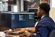 © TrueFrame Collective - Young adult Black man working at desk using dual monitors in modern office, focusing on computer screen while typing on keyboard, short hair visible, professional environment