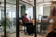 © TrueFrame Collective - Young adult Caucasian man sitting at desk working on computer in modern office, focused expression, glass walls and open workspace visible, colleagues moving in background