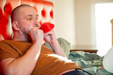Man inflating heart-shaped balloons in a bedroom on a red bed, preparing a romantic setting for Valentine’s Day.