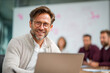 © Daria - Smiling mature businessman with glasses sitting at a laptop in a modern office with colleagues collaborating in the background during a meeting session