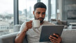 © siraphol - Young man holding coffee mug and tablet on sofa, thoughtful expression in modern living room