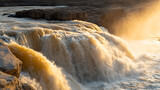 Golden light illuminates a powerful waterfall cascading over rocky ledges. Water rushes with incredible force, creating a mesmerizing display of nature's raw beauty.