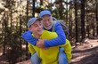 © luciano - Happy senior man enjoying a piggyback ride with a young adult while hiking on a forest trail. Outdoor lifestyle scene showing family bonding, active aging, intergenerational relationship in nature