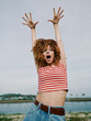 © SHOTPRIME STUDIO - Energetic young woman with curly hair raises arms in excitement, wearing a striped crop top and denim jeans, outdoors by a calm body of water under a bright sky.