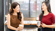 © NAMPIX - Two smiling Asian businesswomen holding coffee cups and chatting in modern office. Casual business meeting and friendly professional relationship in workspace.