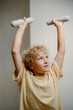 © m-art - A young child with curly hair lifts small weights above their head with focus and determination. The setting is a well-lit room, creating an encouraging atmosphere for exercise.