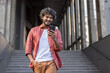 © Tetiana - Indian young man in red shirt standing near building, holding hand in pocket and using mobile phone