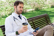 © StockMe - Male doctor in medical uniform with a stethoscope relaxing on a park bench during a coffee break, checking his mobile phone, finding calm and balance outside the hospital. Looking away