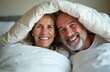 © miss irine - Mature couple laughs under white duvet in bed at home. Happy people share moment of joy and intimacy. Woman and man enjoy cosy morning together, feeling warmth and connection.