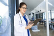 © SOMKID - Doctor woman using digital tablet standing in clinic look at camera, Doctor woman wearing glasses and medical coat working in hospital, Healthcare professional service at modern center.