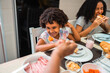 © Nexa - Happy young Black girl smiling while being fed fruit during a family breakfast at home.