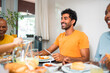 © Nexa - Happy young man in orange t-shirt enjoying a meal with family and friends at home. Diverse group of people sharing breakfast and smiling together at the dining table.