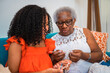 © Nexa - Senior African American grandmother teaching her young adult granddaughter how to crochet while sitting together on a sofa at home.