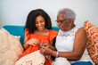 © Nexa - Senior African American grandmother teaching her granddaughter how to crochet while sitting together on a sofa at home.