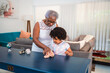 © Nexa - Senior Black woman teaching her granddaughter how to dust a table in a modern living room.