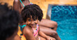 © Nexa - Portrait of a smiling young Black girl with natural curly hair sitting poolside with her family during summer vacation.