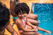© Nexa - Happy African American girl with curly hair sitting on the edge of a swimming pool with her family during summer vacation.