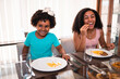 © Nexa - Happy young Black girl and woman laughing while sharing a meal of french fries at home.
