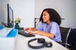 © Nexa - Happy young African American woman with curly hair working or studying on a computer at her home desk, typing on a keyboard with a smile.