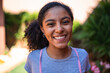 © Nexa - Close-up portrait of a happy young woman with curly hair smiling at the camera in a sunny outdoor garden setting.