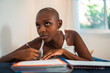 © Nexa - Pensive young African American woman with a shaved head writing in her journal while lying down at home, looking up thoughtfully.