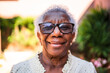 © Nexa - Close-up portrait of a happy senior Black woman with grey hair and glasses smiling at the camera in a sunny garden.