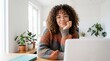© MuzammilZia - Authentic portrait of a happy young woman with curly hair and glasses sitting at her bright home office desk, working remotely on a laptop with cozy plants around her.