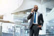 © Prostock-studio - A man in a suit stands in a contemporary office space while talking on his phone. He smiles and appears engaged, with a hand in his pocket. Sunlight enters through large windows.