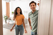 © Prostock-studio - Cheerful young couple stands in the doorway of their modern flat, inviting guests inside with warm smiles. They are excited to host and eagerly await visitors to join them.