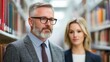 © PStyle - Two professionally dressed people stand between library bookshelves, focused and thoughtful in a scholarly environment.
