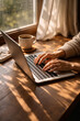 © muhagraph - Person's hands typing on a laptop at a wooden desk with coffee and notebook, bathed in warm sunlight from a window.