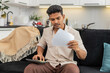 © Andrii Iemelianenko - Indian man at home holds utility bills uses calculator enters numbers for budget and squints at totals. Arabian guy on sofa marks priorities writes notes sets payment order plans monthly costs calmly