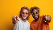 © svastix - Two young men joyfully posing together against a bright yellow backdrop, pointing playfully at the camera in vibrant sunglasses and casual attire, radiating friendship.