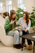 © m-art - Two friends sit together on a plush chair, excitedly sharing and discussing colorful books in a modern shop filled with greenery and soft lighting.
