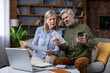 © Liubomir - Senior couple on a sofa, stressed and confused as they compare paper bills with online banking on a smartphone and laptop, struggling with unexpected expenses and budgeting
