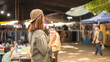 © Happy Photo - A woman stands with a drink in her hand looking at vendors in a busy market. Many people walk around shopping. Tents and stalls surround the area. It is a sunny day
