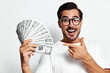 © SHOTPRIME STUDIO - Happy smiling man with glasses holds a fan of cash while pointing at it, showcasing excitement and success, isolated on a plain white background.