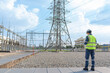 © JD Studio - Electrical engineer in safety vest standing at power substation, monitoring high voltage transmission towers and electricity grid infrastructure for maintenance planning and energy security.