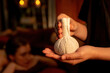 © InfiniteFlow - Hot herbal ball spa massage body treatment with masseur's hand hold or show herb bag at spa. Tranquil and serenity of aromatherapy recreation in warm lighting of candles at spa salon. Quiescent