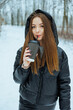 © DSMT - Young woman stands in snowy forest holding drink while wearing warm jacket and enjoying winter outdoors on a cold day