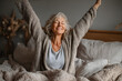 © Daniel - Joyful healthy older woman sitting on bed, waking up after good night rest on comfortable orthopedic mattress, rising up arms and stretching back. Happy mature grandma feeling energetic in morning.