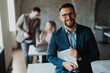 © Novak - Confident male business manager holding a laptop and smiling at the camera with diverse colleagues working in the background of a modern office.